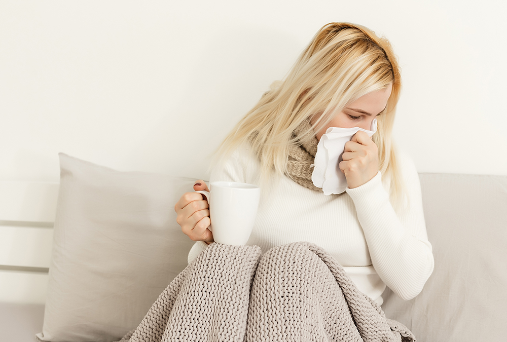 Woman with colds sitting while holding a white cup and blowing her nose